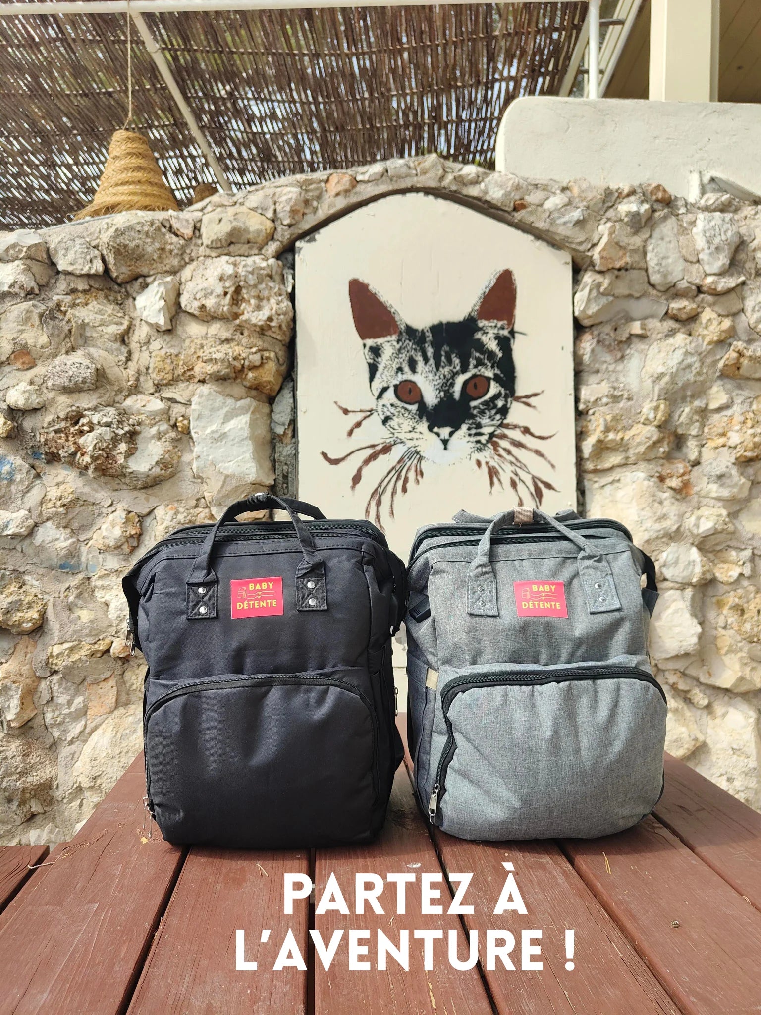 Présentation des deux sacs à langer babydetente couleur noir et couleur gris posés sur une table en bois avec en arrière plan le dessin style street art de la tête d'un chat en grand format sur un mur de pierre - sur la photo le slogan écrit est "Partez à l'aventure"